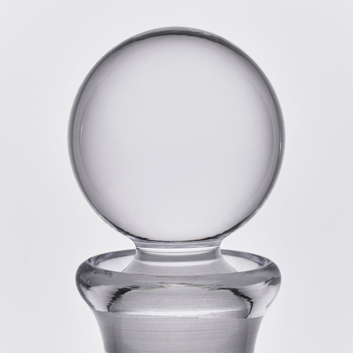 Clear glass decanter with spherical stopper and wide base on neutral background.