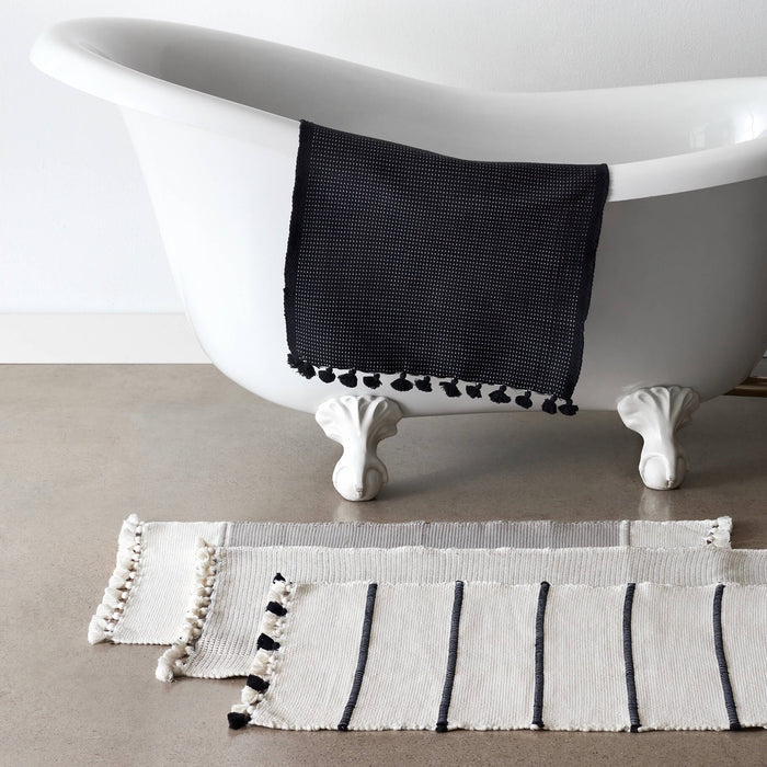 Black bath mat draped over bathtub; three cream bath mats overlaid on floor