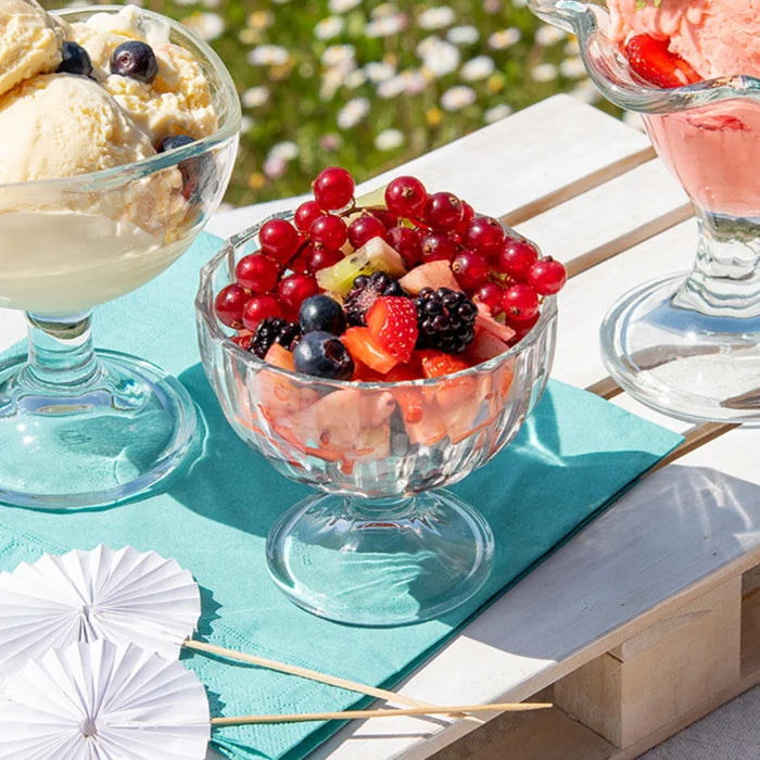 A clear glass pedestal dessert bowl filled with mixed berries and fruit sits on a turquoise napkin on a white wooden table outdoors. Ice cream and a pink dessert are partially visible in adjacent bowls, with blurred greenery in the background.