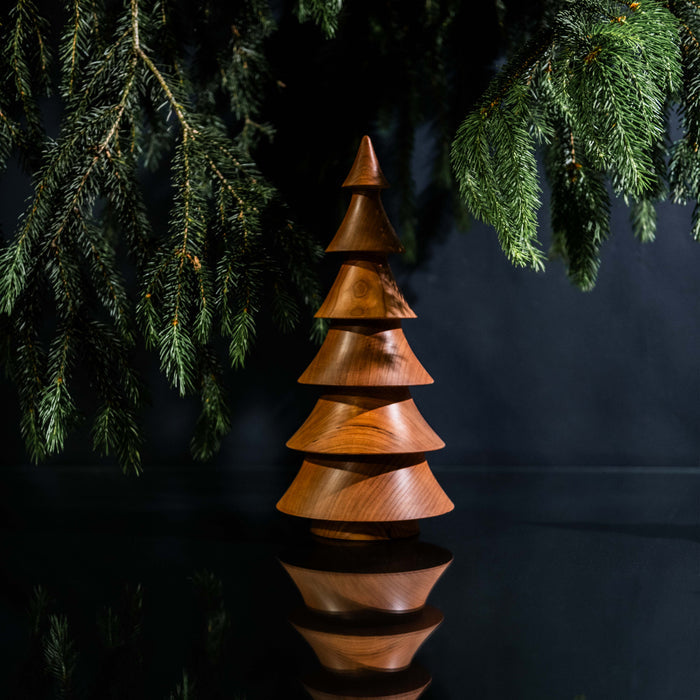 Cherry wood stacked tree decoration with pointed top, surrounded by hanging evergreen branches against dark background.