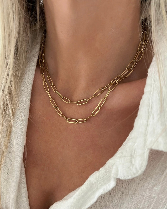 Model wearing two layered gold paperclip chain necklaces of different lengths over a white linen top