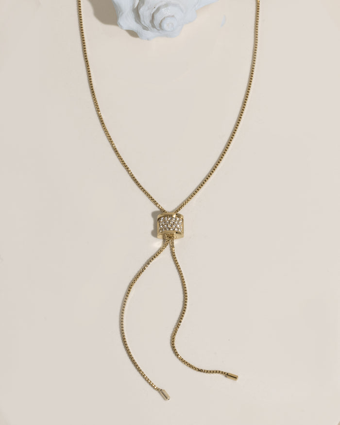 Gold box chain lariat necklace with a crystal-encrusted cylindrical slider bead and dangling chain ends on cream surface