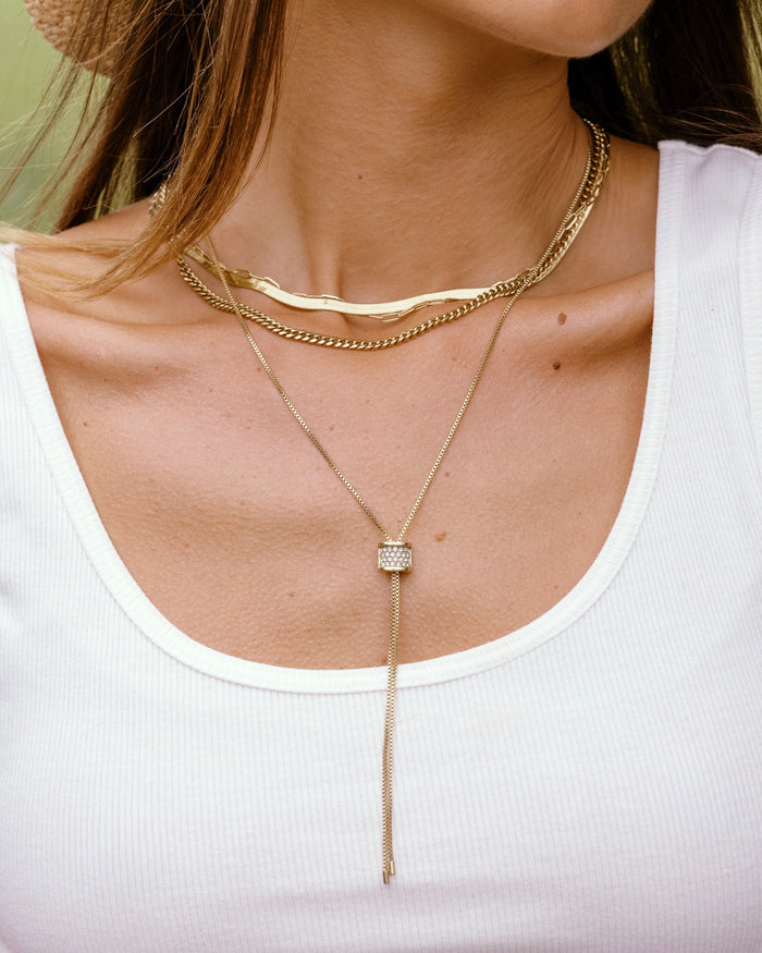 Model wearing three layered gold necklaces: herringbone choker, curb chain, and crystal slider lariat, over a white tank top