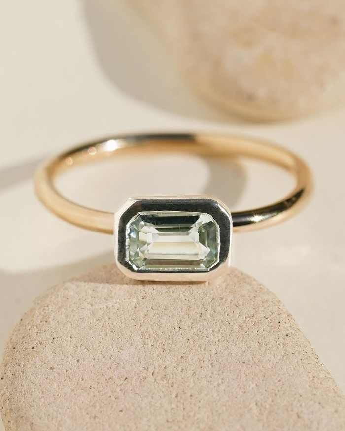 Gold band ring with an emerald-cut light green gemstone in a silver square bezel setting on stone surface