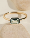 Gold band ring with an emerald-cut light green gemstone in a silver square bezel setting on stone surface