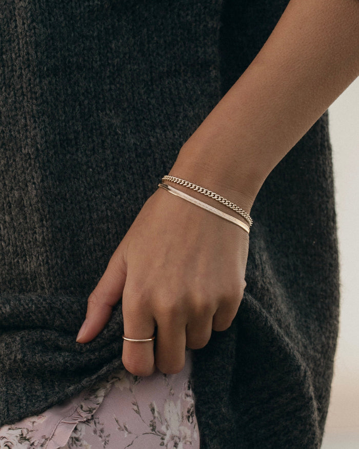 Model wrist wearing stacked gold herringbone bar bracelet and curb chain bracelet with thin gold ring