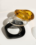 Three stacked resin bangle bracelets in amber tortoise shell, clear, and matte black on white surface with shell