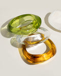 Three stacked transparent resin bangle bracelets in olive green, clear, and amber gold tones on cream surface with shell