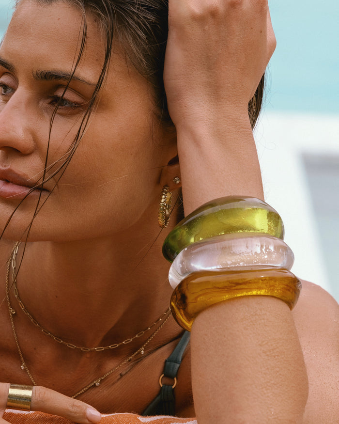 Poolside model wearing three stacked resin bangles in olive, clear, and amber with gold hoop earrings and layered gold necklaces
