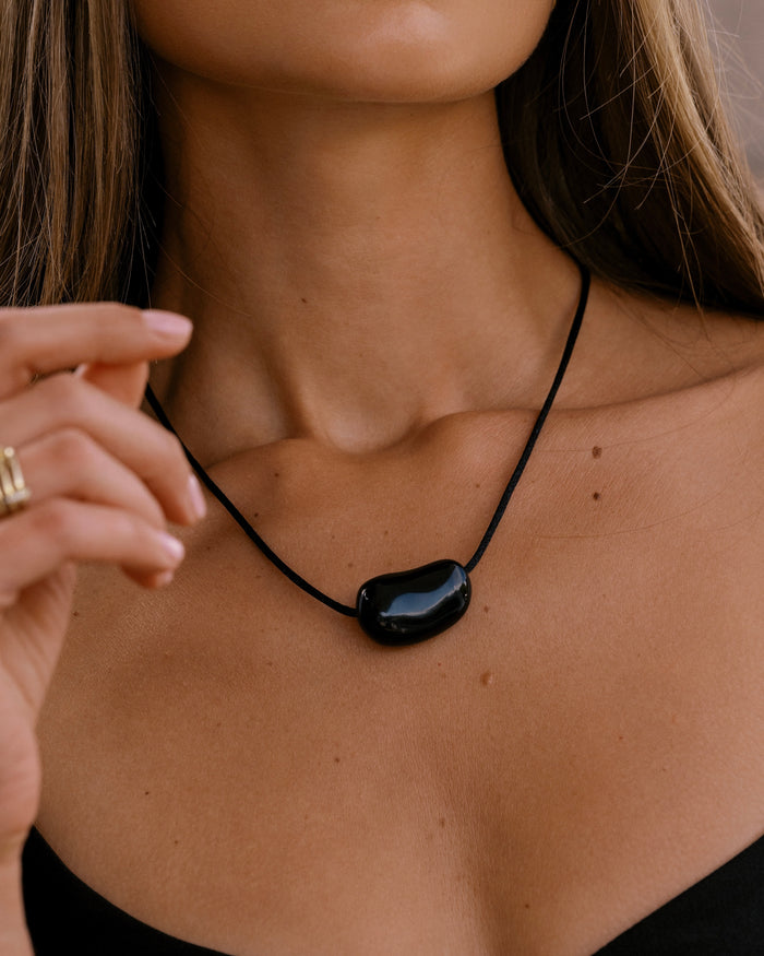 Model wearing a black cord necklace with a polished black organic stone pendant at the neckline with a gold ring