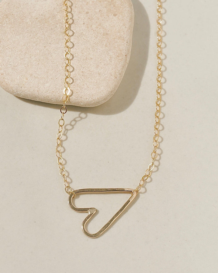 Delicate gold chain necklace with a sideways open-outline gold heart pendant on cream surface with stone pebble
