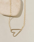 Delicate gold chain necklace with a sideways open-outline gold heart pendant on cream surface with stone pebble