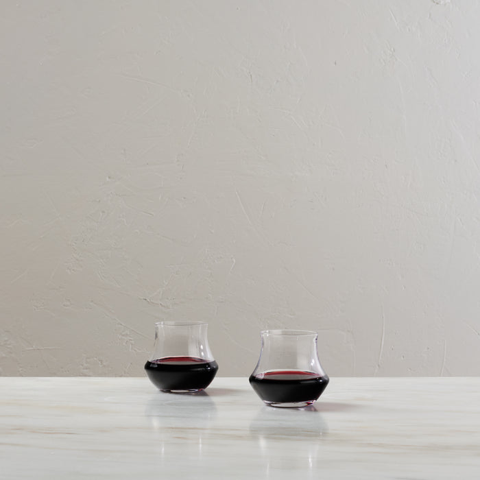 Two clear stemless wine glasses filled with red wine on a white surface.