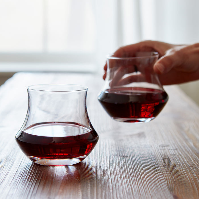 Two clear stemless wine glasses, one filled with red wine and one being held with red wine, on a wooden table.