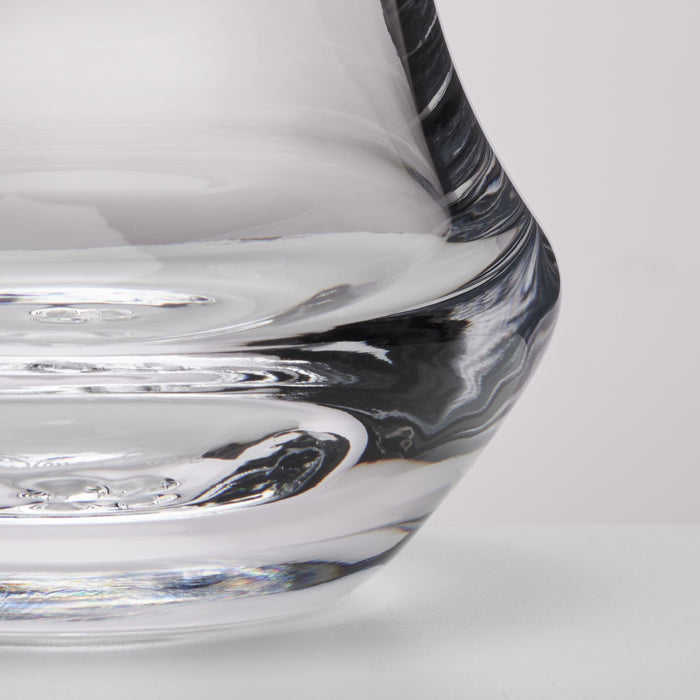 Close-up of a clear glass vessel with a curved, sculptural rim showing reflective glass textures on a light gray background.