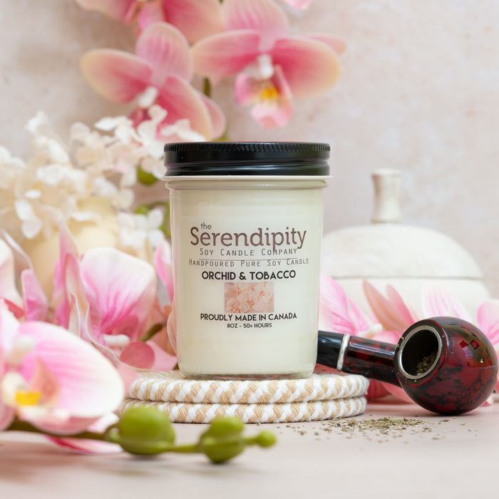 A Serendipity "Orchid & Tobacco" scented candle, positioned in front of some orchids and a pipe