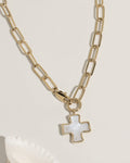 Gold paperclip chain necklace with a mother-of-pearl cross charm pendant on cream surface