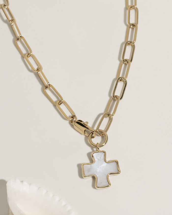 Gold paperclip chain necklace with a mother-of-pearl cross charm pendant on cream surface