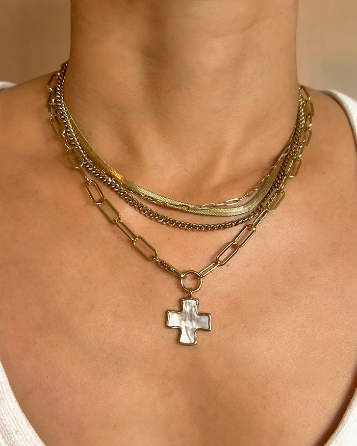 Model wearing three layered gold necklaces including a herringbone choker, curb chain, and paperclip chain with a mother-of-pearl cross pendant