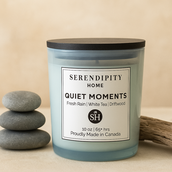 Candle labeled 'Serendipity Home Quiet Moments' with stones and driftwood on a beige background