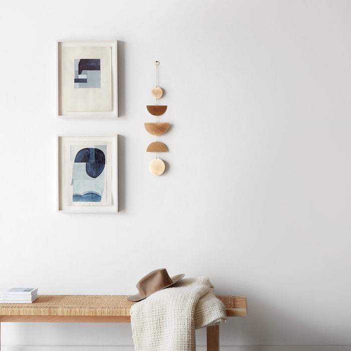 Geometric brass wall hanging in gallery arrangement with abstract art above a woven bench, brass
