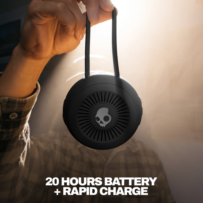 Person holding a black portable speaker with a skull logo, text '20 Hours Battery + Rapid Charge' on a blurred background. | True Black