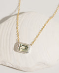 Delicate gold chain necklace with emerald-cut light green gemstone in silver bezel setting, resting on a white shell