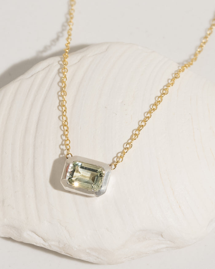 Delicate gold chain necklace with emerald-cut light green gemstone in silver bezel setting, resting on a white shell