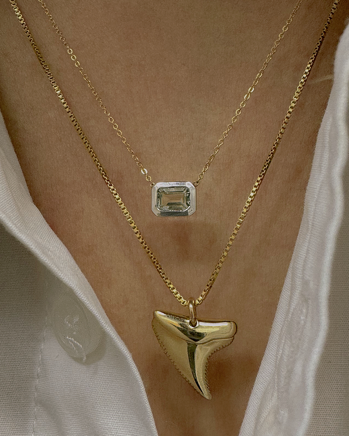 Model wearing two layered necklaces: a gold chain with emerald-cut gemstone pendant and a gold shark tooth charm necklace