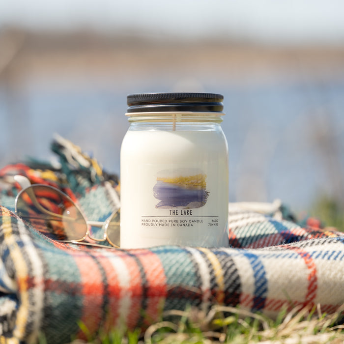 Candle labeled 'The Lake' on a plaid blanket with a blurred natural background
