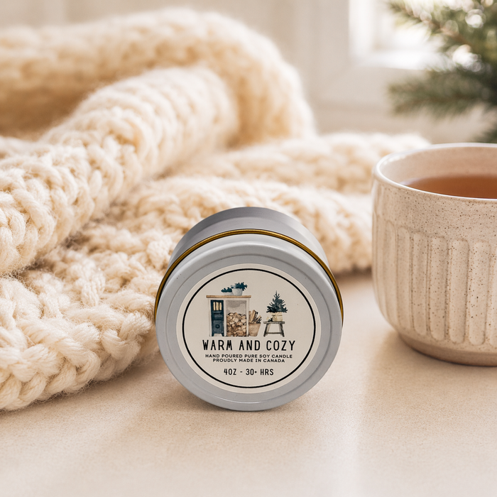 Candle labeled 'Warm and Cozy' next to a knitted blanket and a mug on a light surface.